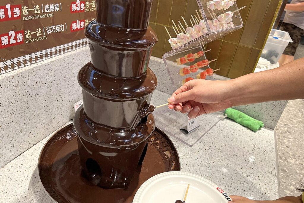 Großer Schokoladenbrunnen auf einem Buffet, ein Gast hält einen Spieß in den Schokoladenstrom