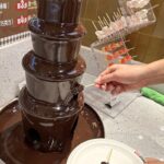 Großer Schokoladenbrunnen auf einem Buffet, ein Gast hält einen Spieß in den Schokoladenstrom